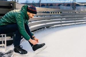 People, sport, winter, leisure concept. Smiling delightful male laces up skates, going to practice on ice ring, being in good mood, likes winter and go skating with best friends. Hobby concept photo