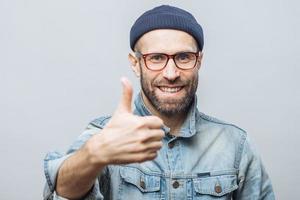 Happy delighted man with stubble raises thumb as shows his approval, dressed in stylish clothing, isolated over white studio background. Satisfied middle aged male gestures indoor. Body language photo