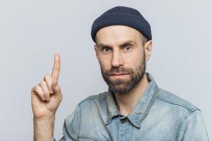 Indoor shot of handsome man with serious look, has pleasant appearance, thick beard with mustache, wears fashionable clothing, raises fore figer as gets brilliant idea, isolated over grey background photo