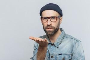 Pleasant looking young bearded male with mustache, wears fashionable glasses, black hat and denim shirt, blows air kiss at camera, expresses his love to girlfriend, isolated over grey background photo