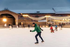 un alegre hombre barbudo pasa la Navidad en una majestuosa pista de hielo decorada con luces, patina sobre hielo, se divierte, disfruta de su pasatiempo y del clima nevado del invierno. gente, ocio, concepto de estilo de vida activo foto