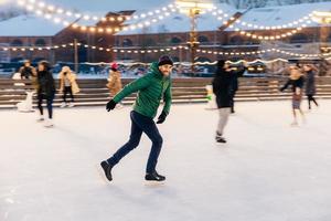 toma al aire libre de un hombre atractivo con barba, usa ropa de invierno abrigada, practica patinaje sobre hielo decorado con luces, tiene una expresión alegre, disfruta de su acción favorita foto