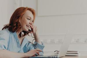 Profile shot of happy redhead woman laughs positively, has online communication with friend, takes break after work, wears blue shirt, poses in coworking space. People, freelance work, distance job photo