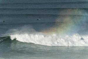 Big giant waves crashing in Nazare, Portugal creating a rainbow in the ocean. Surfers and jet skis in the water. Biggest waves in the world. Touristic destination for surfing. photo