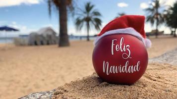 bomba de navidad en el sombrero de santa con palabras feliz navidad en español en la playa tirada en la arena con palmeras y cielo azul en el fondo. feliz navidad desde el paraíso, isla exótica. foto