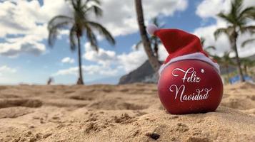 Christmas bomb in Santa's hat with words Merry Christmas in spanish on the beach lying on the sand with palm trees and blue sky on the background. Merry Christmas from paradise, exotic island. photo