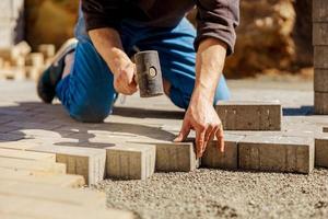 Young man laying gray concrete paving slabs in house courtyard on gravel foundation base. Master lays paving stones. Garden brick pathway paving by professional paver worker. Repairing sidewalk. photo