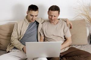 feliz pareja gay con ropa informal pasando tiempo juntos en casa y viendo películas en la laptop. dos hombres caucásicos relajándose. Relaciones homosexuales y amor alternativo. interior acogedor. foto