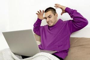 Angry and dissatisfied man with violet hoodie sitting on bed with laptop computer. Man using notebook to surf in internet, read news, watch movie, study or work online. Negative emotions, stress. photo