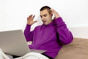 Young caucasian man with violet hoodie sitting on bed with laptop computer. Man using notebook to surf in internet, read news, watch movie, study. Surprised, positive emotions. Won a prize online. photo