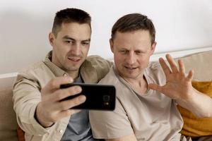 Happy gay couple with casual clothes holding smartphone and talking with friends online, having video call. Homosexual relationships and alternative love. photo