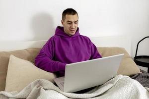 Young caucasian man with violet hoodie sitting on bed and holding laptop computer. Man using notebook to surf in internet, read news, watch movie, study or work online, call friends. Relaxing at home. photo