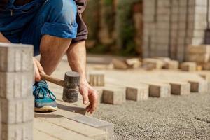 Young man laying gray concrete paving slabs in house courtyard on gravel foundation base. Master lays paving stones. Garden brick pathway paving by professional paver worker. Repairing sidewalk. photo