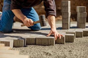 Young man laying gray concrete paving slabs in house courtyard on gravel foundation base. Master lays paving stones. Garden brick pathway paving by professional paver worker. Repairing sidewalk. photo