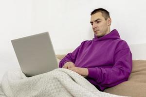 Young caucasian man with violet hoodie sitting on bed and holding laptop computer. Man using notebook to surf in internet, read news, watch movie, study or work online. Relaxing at home. photo