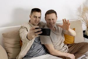 feliz pareja gay con ropa informal sosteniendo un teléfono inteligente y hablando con amigos en línea, haciendo videollamadas. Relaciones homosexuales y amor alternativo. foto