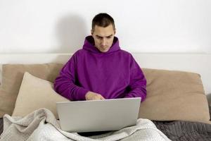 Young caucasian man with violet hoodie sitting on bed and holding laptop computer. Man using notebook to surf in internet, read news, watch movie, study or work online. Relaxing at home. photo