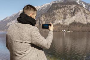 hombre adulto joven tomando fotos con su teléfono inteligente, disfrutando de las montañas, el lago, el buen tiempo, el cielo azul, el sol. hermoso, increíble paisaje. turismo, vacaciones, vacaciones, tiempo de viaje. turista haciendo foto.