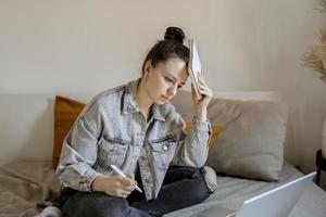 Young beautiful woman with casual clothes sitting on the bed at home with laptop computer and studying. Girl is sad and tired. Negative emotions, stress, mental problems, deadline. Distance education. photo