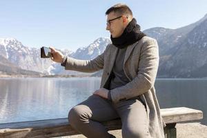 Young adult man sitting outdoors on bench and making selfie with his smartphone, enjoying mountains, lake, good weather, blue sky, sun. Beautiful, amazing landscape. Holiday, vacation, travel time. photo