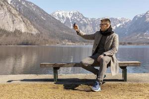 Young adult man sitting outdoors on bench and making selfie with his smartphone, enjoying mountains, lake, good weather, blue sky, sun. Beautiful, amazing landscape. Holiday, vacation, travel time. photo