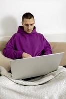 Young caucasian man with violet hoodie sitting on bed and holding laptop computer. Man using notebook to surf in internet, read news, watch movie, study or work online. Relaxing at home. photo