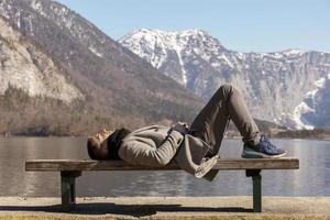 Young man lying outdoors on bench and enjoying mountains, snow, good weather, blue sky, sun. Beautiful landscape. Time with yourself, dreaming, relaxation, mental health. Tourism, holiday, travel. photo