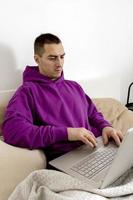 Young caucasian man with violet hoodie sitting on bed and holding laptop computer. Man using notebook to surf in internet, read news, watch movie, study or work online, call friends. Relaxing at home. photo