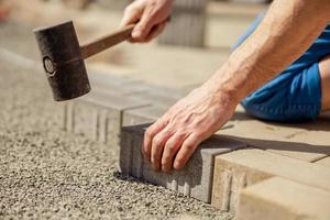 Young man laying gray concrete paving slabs in house courtyard on gravel foundation base. Master lays paving stones. Garden brick pathway paving by professional paver worker. Repairing sidewalk. photo