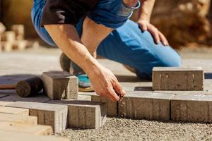 Young man laying gray concrete paving slabs in house courtyard on gravel foundation base. Master lays paving stones. Garden brick pathway paving by professional paver worker. Repairing sidewalk. photo