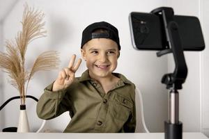 adorable, lindo, pequeño bloguero grabando un blog de estilo de vida, hablando con la cámara del teléfono inteligente en un trípode. joven influencer filmando vlog para su canal. niño hace video para sus seguidores en línea. foto