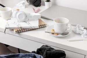 Messy table with laptop, dirty cups, and stationary items. Cluttered workplace, desk. Concept of being overwhelmed by working or studying. Mess and chaos at home. Deadline, stress. photo