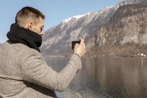 Young adult man taking picture with his smartphone, enjoying mountains, lake, good weather, blue sky, sun. Beautiful, amazing landscape. Tourism, holiday, vacation, travel time. Tourist making photo. photo