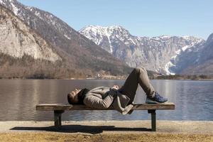 Young man lying outdoors on bench and enjoying mountains, snow, good weather, blue sky, sun. Beautiful landscape. Time with yourself, dreaming, relaxation, mental health. Tourism, holiday, travel. photo