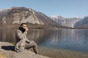 Young adult man sitting outdoors, drinking coffee and enjoying mountains, lake, good weather, blue sky and sun. Beautiful landscape. Time with yourself, dreaming, relaxation, mental health. Holiday. photo