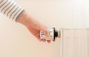 Woman regulates temperature at home with the heating thermostat to save energy, close-up with hand. Temperature control valve on radiator. White heater on wall with curtain in modern apartment. photo