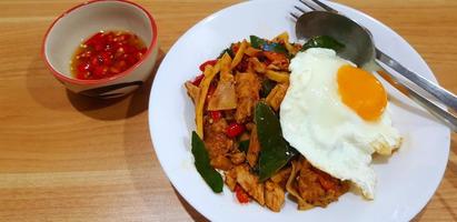 Stir fried chicken with sour bamboo shoot, red chili, kaffir lime leaves and fried egg in white plate or dish and spicy fish sauce on wooden table or wood background. Asian food with spoon and fork. photo