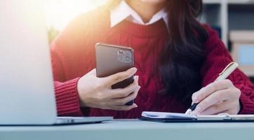 Young business woman sitting in office at table and using smartphone. On desk is laptop and tablet computer, on screen charts and graphs. Woman analyzing data. Student learning online. photo