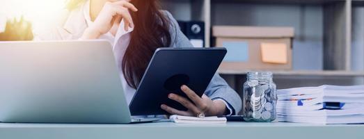 Young business woman sitting in office at table and using smartphone. On desk is laptop and tablet computer, on screen charts and graphs. Woman analyzing data. Student learning online. photo