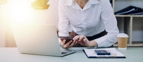 Young business woman sitting in office at table and using smartphone. On desk is laptop and tablet computer, on screen charts and graphs. Woman analyzing data. Student learning online. photo