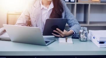 Young business woman sitting in office at table and using smartphone. On desk is laptop and tablet computer, on screen charts and graphs. Woman analyzing data. Student learning online. photo