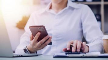 Young business woman sitting in office at table and using smartphone. On desk is laptop and tablet computer, on screen charts and graphs. Woman analyzing data. Student learning online. photo
