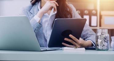Young business woman sitting in office at table and using smartphone. On desk is laptop and tablet computer, on screen charts and graphs. Woman analyzing data. Student learning online. photo