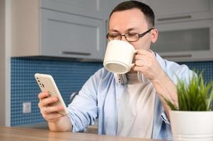 Young Asian man wearing galsses sitting and using smartphone while drinking coffee at home. Modern technologies concept.Online cimmunication photo