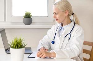 retrato de una joven y confiada doctora caucásica con uniforme médico blanco sentada en el escritorio llenando los formularios mirando la pantalla del portátil foto