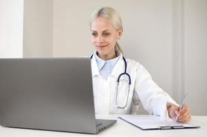 retrato de una joven y confiada doctora caucásica con uniforme médico blanco sentada en el escritorio llenando los formularios mirando la pantalla del portátil foto