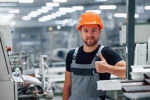 Shows thumb up. Well done. Everything is good. Industrial worker indoors in factory. Young technician with orange hard hat photo