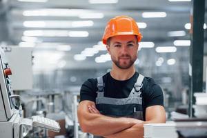 Stands with arms crossed. Portrait of male industrial worker indoors in factory. Young technician with orange hard hat photo