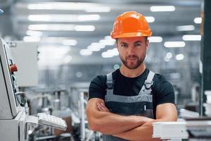 Stands with arms crossed. Portrait of male industrial worker indoors in factory. Young technician with orange hard hat photo