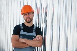 Leaning on the wall. Serious industrial worker indoors in factory. Young technician with orange hard hat photo